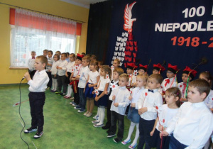 Chłopiec mówi do mikrofonu. Z tyłu dzieci ustawione w szeregu.