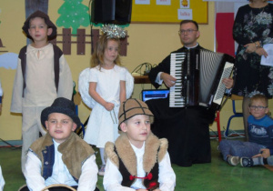 Czworo dzieci w strojach jasełkowych. Za nimi ksiądz gra na akordeonie.
