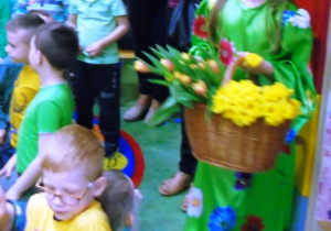 Dziewczynka w zielonej sukience trzyma koszyk z kwiatami.