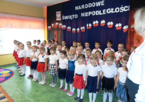 Grupa dzieci odświętnie ubranych stoi na baczność. W tle napis " Narodowe święto niepodległości".