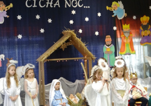 Dziewczynka w stroju anioła mówi wiersz, obok niej stoi dziewczynka również w stroju anioła.W tle widac napis " Cicha noc".