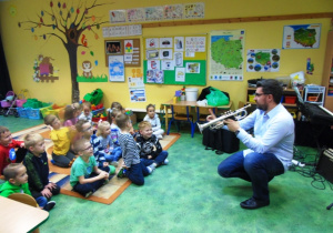 Grupa dzieci siedzi na dywanie.Muzyk z trąbką kuca przed dziećmi i pokazuje im instrument.