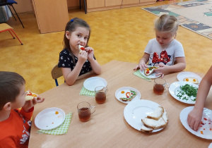Dzieci siedza przy stolikach i jedzą kanapki.