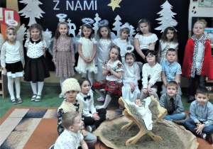 Dzieci ubrane odświętnie stoją, za nimi napis Kolędujcie z nami.