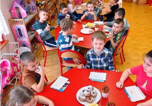 Dzieci siedzą przy stoliku i jedzą pączki.