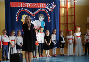 Dzieci ubrane odświętnie stoją w półkolu na scenie, za nimi dekoracja na Święto Niepodległości. Przed dziećmi stoi kobieta i przemawia przez mikrofon.