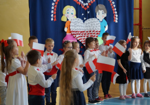 Występujące przedszkolaki śpiewają piosenkę i trzymają chorągiewki.