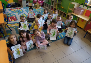 Dzieci siedzą na dywanie i prezentują swoje prace.