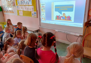 Grupa dzieci siedzi przed tablica interaktywną.