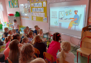 Grupa dzieci siedzi przed tablica interaktywną i oglądają prezentację multimedialną.