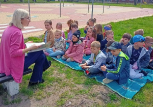 Dzieci siedzą na kocu na trawie i słuchają książki czytanej przez nauczyciela.