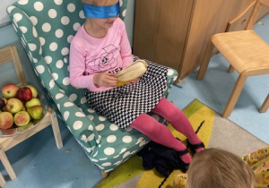 Na zdjęciu widać dziewczynkę która siedzi na fotelu i ma zawiązane oczy i trzyma w dłonicach banana