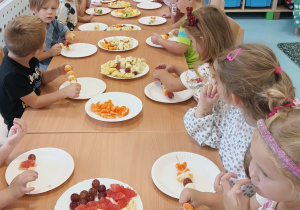 Grupa dzieci siedzi przy stoliku i zajada owocowe szaszłyki