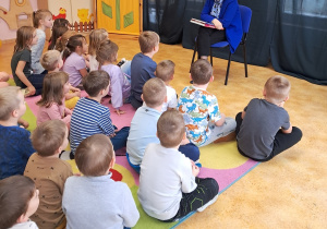 Dzieci siedzą na dywanie i słuchają wiersza czytanego przez zaproszonego gościa.
