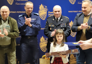 Na zdjęciu widać czterech panów w tym dwóch policjantów policjantów przed nimi stoi dziewczynka która ma wręczaną nagrodę