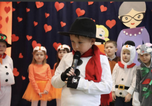 Na zdjęciu chłopiec w czarnym kapeluszu trzyma mikrofon. Za nim widać występujące dzieci.