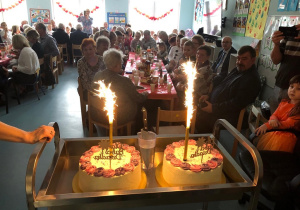 Na zdjęciu są dwa torty z odpalonymi racami. W tle widać dzieci ze swoimi dziadkami.
