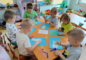 Przy stoliku siedzą chłopcy i przyklejają kolorowe paski bibuły.