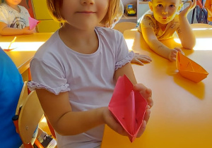 Dziewczynka i chłopiec siedza przy stoliku i pokazują zrobione przez siebie łódki metodą origami.
