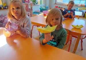 Dziewczynka i chłopiec siedza przy stoliku i pokazują zrobione przez siebie łódki metodą origami.