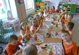 Grupa przedszkolaków siedząca przy stolikach w czasie słodkiego poczęstunku, trzymająca w rękach wykonane papierowe marchewki