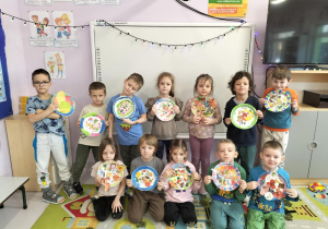 Grupa przedszkolaków prezentująca ozdobione przez siebie papierowe talerzyki ze zdrowymi produktami.
