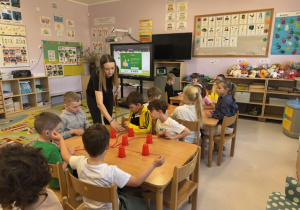 Przedszkolaki siedzące przy stolikach w trakcie gry zręcznościowej wraz z nauczycielką.
