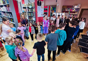 Przedszkolaki wraz z nauczycielką i uczniami w kostiumach stojący w kole i trzymający się za ręce.