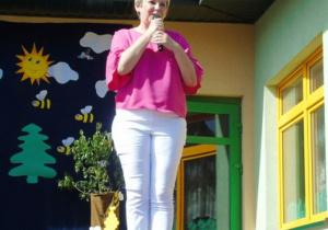 Kobieta w różowej bluzce i białych spodniach przamawia przez mikrofon