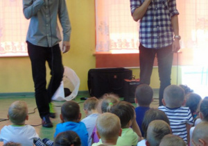 Dwaj mężczyźni stoją i świewająprzez mikrofon przed grupą dzieci