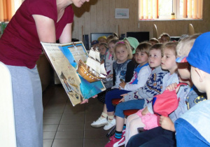 Dzieci siedzą a kobieta stoi przed nimi i pokazume i książkę 3D