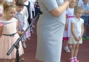 Kobieta przemawia przez mikrofon za sią stoi duża grupa dzieci