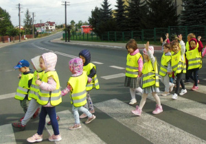 Dzieci w kamizelkach z podniesiona jedną ręka ida przez przejście dla pieszych.