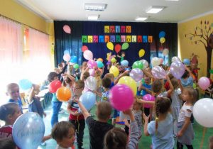 Dzieci tańczą i trzymaja balony. W tle na granatowym tle napis" Dzień przedszkolaka".