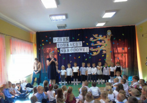 Dzieci odświętnie ubrane stoją ustawione pod napisem " Dzień edukacji narodowej".