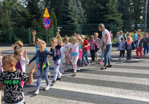 Dzieci przechodzą przez przejście dla pieszych. Mają podniesioną jedną rękę.