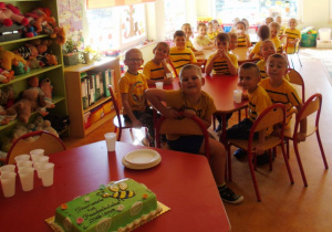 Dzieci w żółtych koszulkach z czarnymi paskami siedzą przy stoliczku obok nich na stoliku leży zielony tort leży