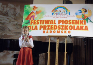 Dziewczynka śpiewa na scenie. W tle napis: Festiwal piosenki dla przedszkolaka.