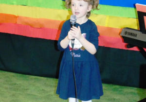 Dziewczynka w niebieskiej sukience mówi do mikrofonu.