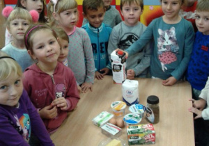 Dzieci stoją przy stoliku. Na stoliku leżą produkty mleczne.