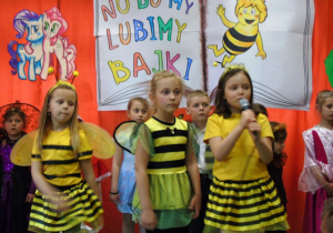 Trzy dziewczynki w strojach pszczółe. Jedna z nich śpiewa do mikrofonu.