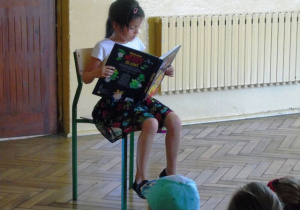 Dziewczynka siedzi na krześle i czyta książkę.