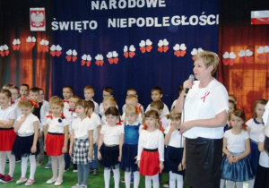 Dzieci odświętnie ubrane stoją w trzech rzędach. Przed nimi stoi kobieta i mówi do mikrofonu.
