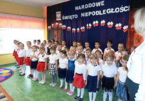 Grupa dzieci odświętnie ubranych stoi na baczność. W tle napis " Narodowe święto niepodległości".