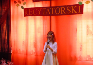 Dziewczynka w białej sukience stoi na podwyższeniu i mówi wiersz do mikrofonu.