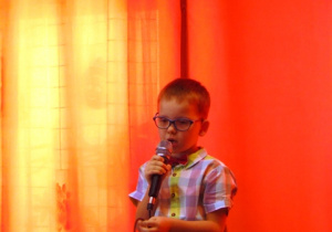 Chłopiec w okularach mówi wiersz do mikrofonu. W tle pomarańczowa zasłona.