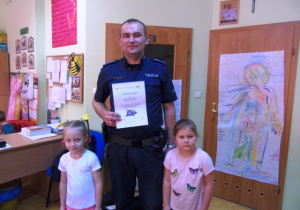 Policjant stoi i trzyma podziękowania. Obok niego stoją dwie dziewczynki.