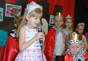 Dziewczynka w sukience i koronie na głowie trzyma mikrofon i mówi wiersz.