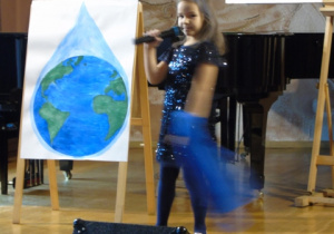 Dziewcznka stoi na scenie i śpiewa do mikrofonu.W ręku trzyma niebieską chustkę. Obok niej na sztaludze stoi obrazem a na nim narysowana Ziemia.