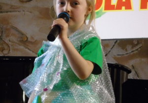 Dziewczynka w zieloym ubraniu stoi na scenie i śpiewa do mikrofonu.
